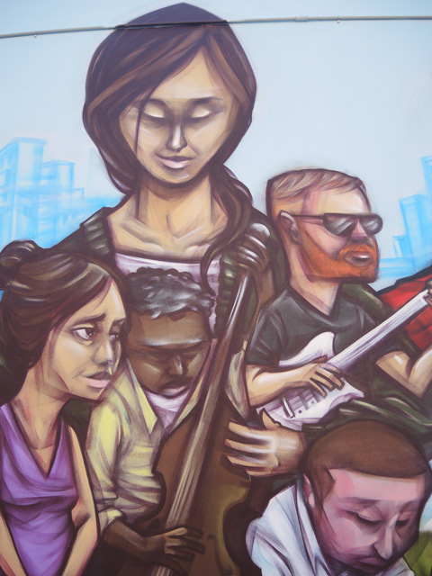 part of a mural by elicser of people - 4 people, a woman overlooks them, a small black man on a cello and a man with beard and glasses on a white guitar. 