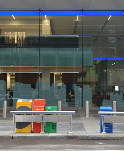 newspaper boxes in different colours in front of a large window of the BMO building, a city scape is etched into the window. Some reflections in the window too 