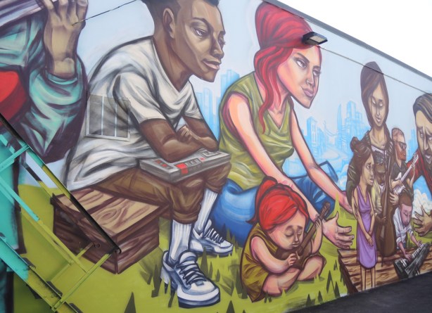 mural, part of, by elicser, people sitting outside, a young child with red hair sits on the grass, her mother behind her. A black man sits on a wooden seat, a cassette player on his lap