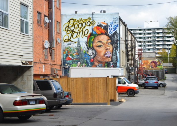 view looking down an alley, cars parked behind buildings, two murals, Welcome to Reggae Lane, 