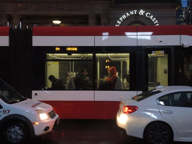 two white cars in front of a stopped streetcar, 514 Cherry, new streetcar, in front of the Elephant and Castle bar on King Street, people sitting on the streetcar are visible, dark outside, wet and rainy evening