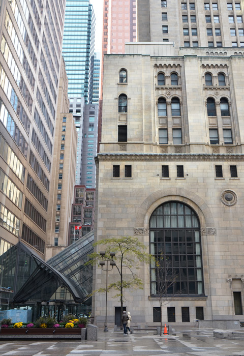 two people walk across a courtyard behind an old stone building that is surrounded by newer glass and steel high rises. 