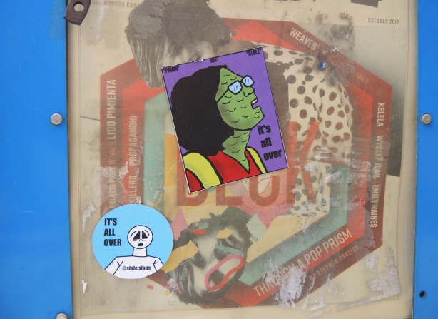 two it's all over stickers on a poster on a wall. one sitcker is a blue circle and the other is a girl with a green face and glasses looking towards the sky 