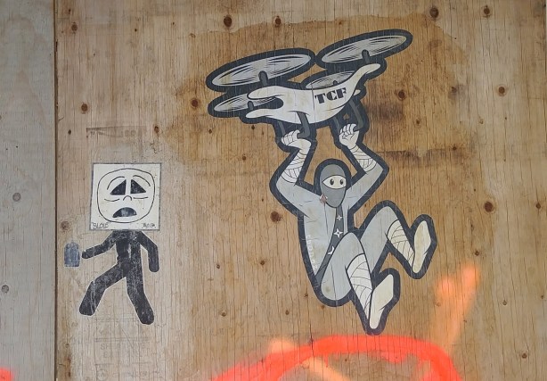 two pasteups on a plywood board on hoardings, one is an urban ninja squadron character holding onto a drone in flight and the other is a simplistic man with a frowny face, an its all over character, both are in black and whtie 