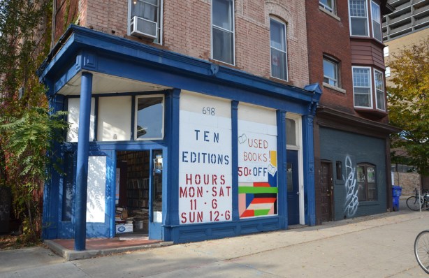 Ten Editions, a used book store on a corner, blue trim, large windows covered with white paper, door is open, stack of boos can be seen inside, old brick building