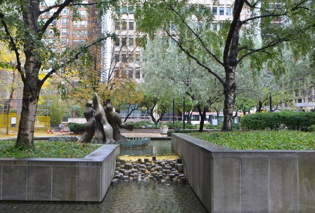 a small waterfall, over rocks, between two concrete fence around grassy areas, trees, small park 
