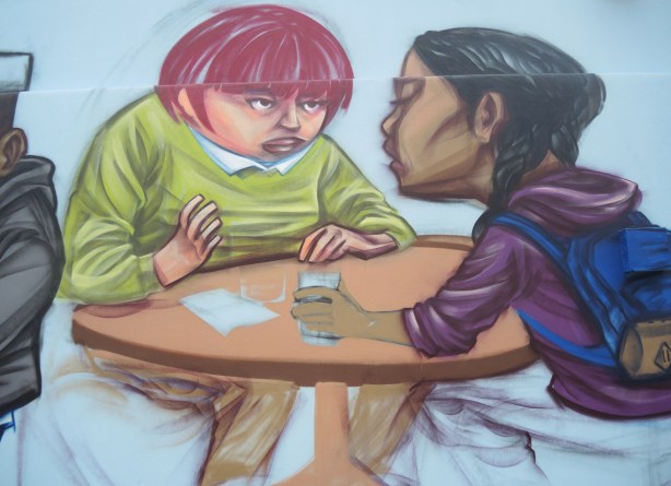 part of a partially completed mural, two girls sitting at a table and talking, one with brown skin and long black hair, one with short red hair, a glass of water in one hand 