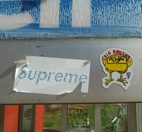 two stickers on the side of Bell telephone booth one just has the word supreme and the other is an uber 5000 yellow birdie holding two words in red letters, 