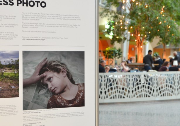 part of a world press photo display at Brookfield Place, with a picture of sick young girl, in the background, people are sitting in a cafe 