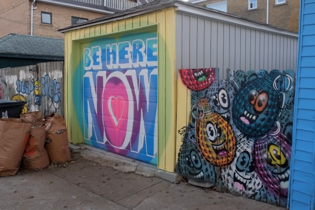 a garage door with the words Be Here Now beside a fence painted with spud bombs, hand grenade shaped characters with eyes and smiling mouths
