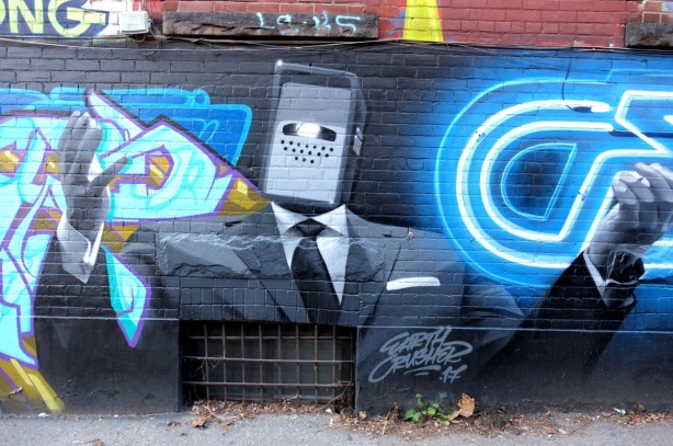 mural in Graffiti Alley by earth crusher - man in black suit, white shirt and black tie, small rectangle machine head and a smart phone in each hand 