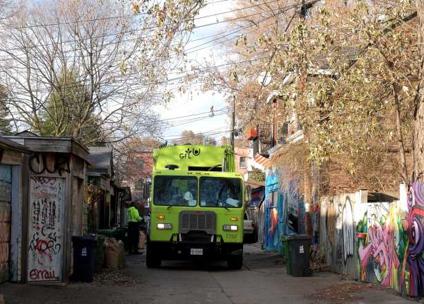 a green GFL garbage truck in a lane as men pick up garbage on garbage day
