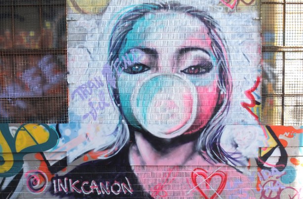 street art painting by inkcanon of a woman blowing a large bubble with bubble gum, in shades of pink and blue 
