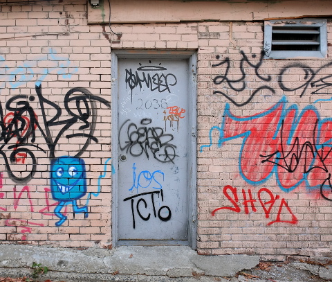 pink brick building with grey metal door, lots of scrawl graffiti on the walls, one litle blue man roughly drawn by the door 