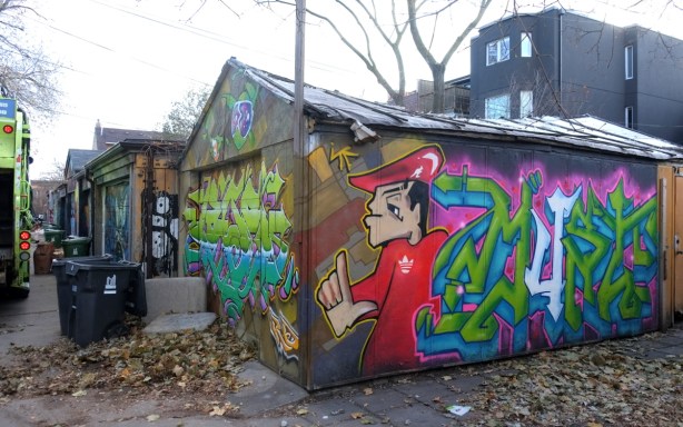 garage with two sides painted in street art, lane, backs of houses, no leaves on tree,