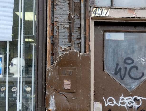 exterior wall, brown paint, number of 437 above the door, cracked wood plywood beside the door, old and broken intercom system that has been painted over, next door window is a store, with mannequin head on a shelf 