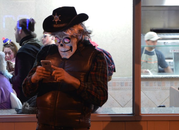a man wearing a cowboy hat with a sheriff's bagde, and a skull mask checks his phone while standing outside pizza pizza, window lit up and people inside including a man making pizza 