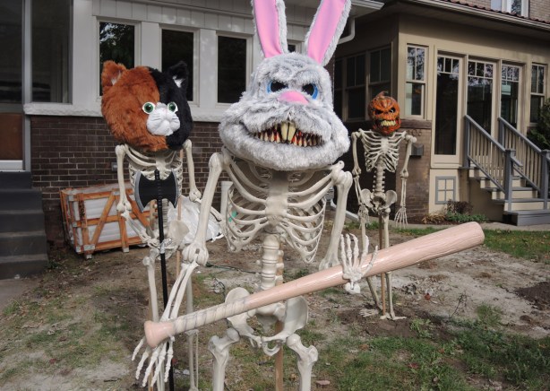 three life sized skeletons in a front yard, a rabbit mask on one which is also holding a baseball bat, a pumpkin head, and one with a bear mask that is half brown and half black 