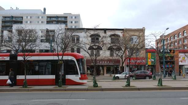 a new ttc streetcar on Spadina, down the middle of the street, with young trees growing along side the tracks, old brck building in the background, some cars, 