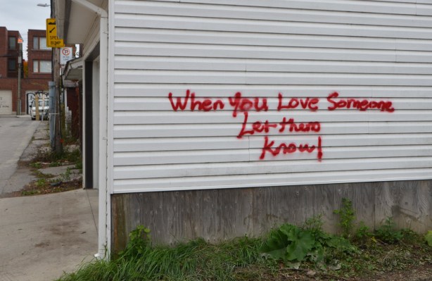 red words painted on the side of a white building in an alley, words say - When you love someone, let them know 