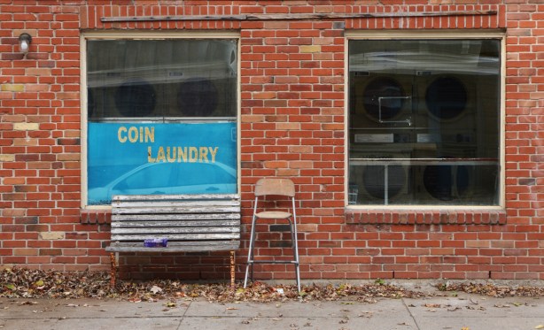 front of a laundromat. blue sign that says coin laundry, an old bench and an old chair sitting outside by the front door. two windows through which you can see the washing machines 