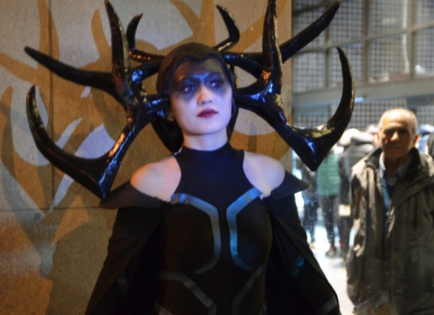 woman in black with large black antlers stand against a wall, an older man is looking at her 