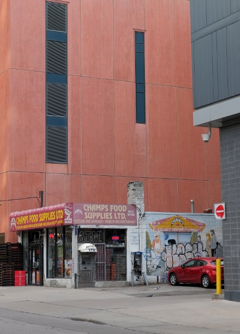 older small building Champs Food Suplies Ltd with a large new condo behind it, red car parked beside it, mural on the side wall 