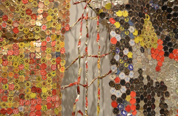 artwork hung on a wall, covers the wall, by El Anatsui, a Ghanese artist, made of different colours of liquor bottle caps, metal, close up