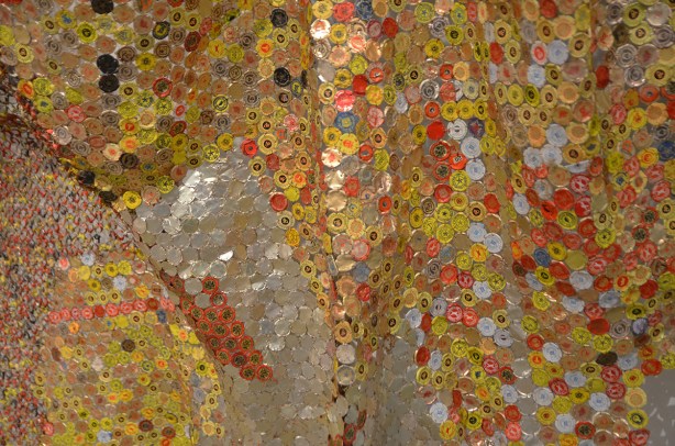 artwork hung on a wall, covers the wall, by El Anatsui, a Ghanese artist, made of different colours of liquor bottle caps, metal, close up