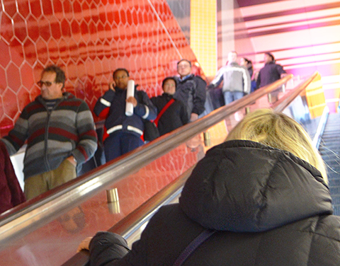 people on an escalator, red hexagonal tiles on the wall beside them, lots of streaks of pink and yellow light above them at the top of the escalator, Finch West subway station TTC, toronto