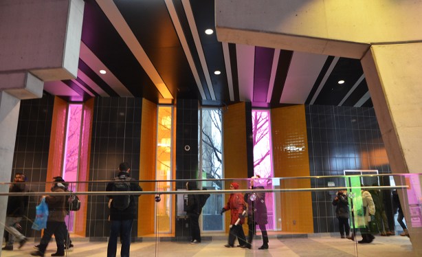 people passing through Finch West station with its tall vertical windows covered in colours, pink, blue and yellow, also with its stripes of white and dark grey tiles