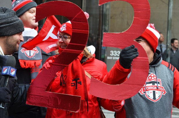 a reporter holding a microphone, interviewing a couple who are watching the Tornto FC parade, she is holding a large red 2 and he is holding a large red 3, the number 23, They are wearing tfc hats andscarves and sweatshirts