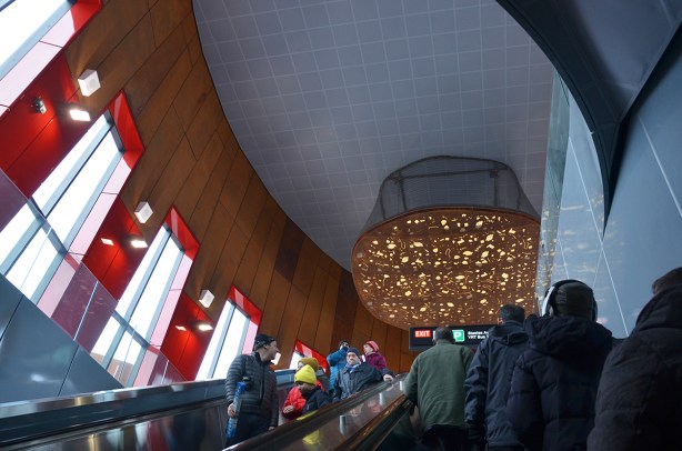 interior of Pioneer Village subway station, top of one of the escalators, vertical windows looking outside, some red glass as accents, a large light artwork on the ceiling, people on the escalators