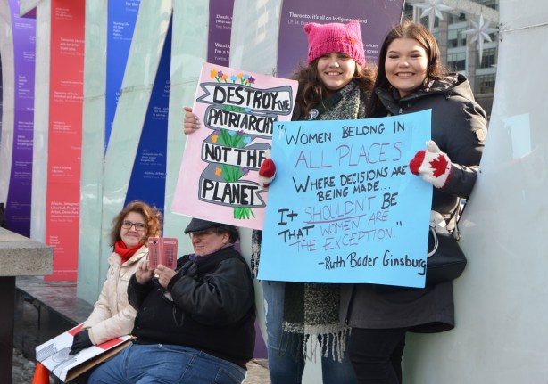 Womens March, Nathan Phillips square, two young women sitting in the O of the 3D Toronto sign, each with a sign, a couple behind them - the woman looking at them, the man taking a picture with his phone. The cover on his phone is a British callbox (phone booth)