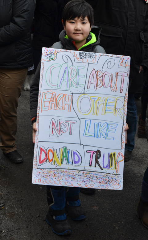 boy with a large hand drawn sign on white bristol board that says care about each other not like donald trump