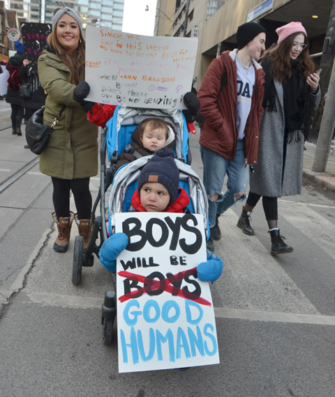 Womens march, protest parade - mother pushing double stroller, with two boys in in, sign that says boys will be good humans, 