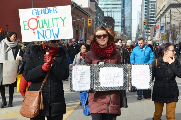 Toronto womens march and protest, a woman with a sign that says gender equality now, and a woman with a sign that says end violence against women along with the names of women who have been killed by their partners. 