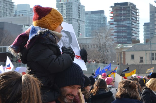 young girl on her father's shoulders, wearing hat and scarf and holding a reistence sign with a picture of Princess Leia on it. 