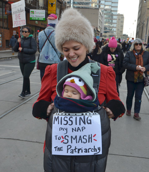 mother and daughter, Womens march, protest parade, daughter is a baby in a front carrier, sign ver her says Missing my nap to smash the patriarchy 
