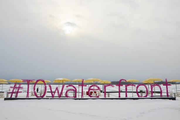 H 2 O park on Toronto's waterfront with yellow umbrellas over white Muskoka chairs on what is a beach in the summer but is covered with snow in the picture. A pink sign that says #TOwaterfront made of pink wood that is supposed to look like pieces of driftwood 