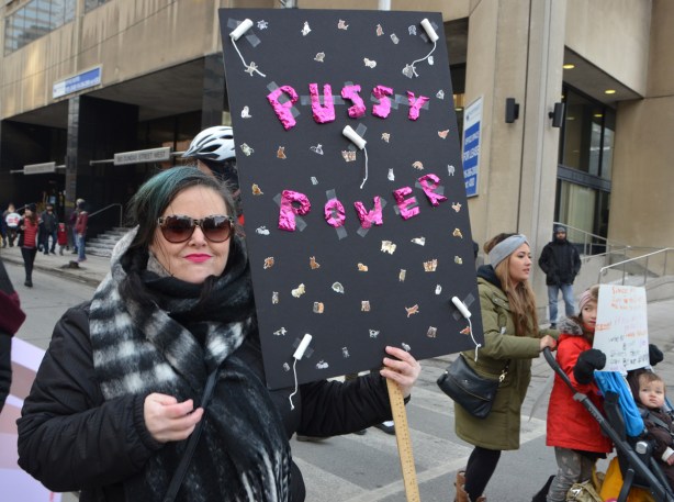 Womens march, protest parade - a woman holding a black placard with pussy power in pink, stickers of cats, and some tampons on it 