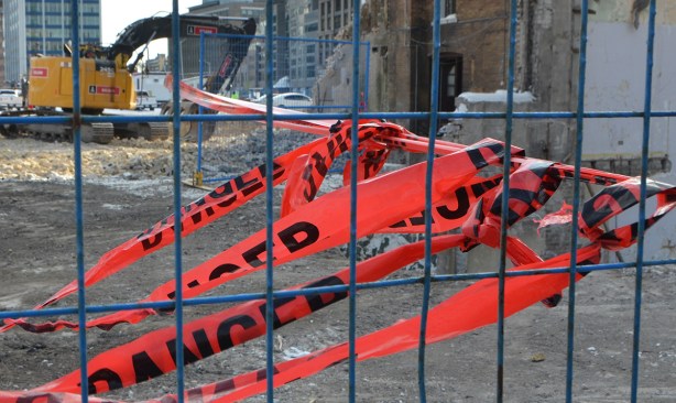 red plastic danger tape blows in the wind. one end is tied to a blue fence and one strand is also tied to a building being demolished 