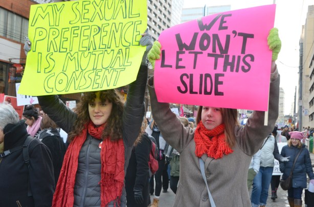 two women holding up signs, a pink one that says we won't let this one slide and a yellow sign that says my sexual preference is mutual consent 