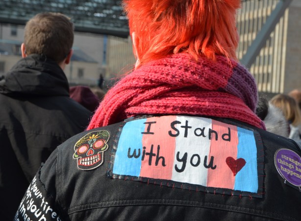 Womens March, Nathan Phillips square, from the back, a woman with short and very orange hair, has a patch on the back of her black jacket, rainbow flag with I Stand with you