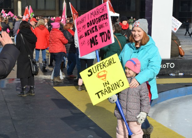 Womens March, Nathan Phillips square, by the rink a mother and daughter getting their picture taken. Both are holding signs, the daughter's sign is yellow and says Supergirl in training, mother's sign is pink and says 