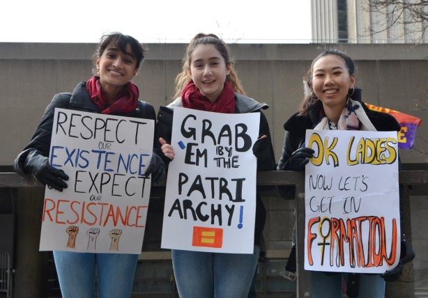 Womens March, Nathan Phillips square, 3 girls watch the protest march go by, they are each holding a sign - Respect my existence or expect resistance, the second is Grab 'em by the Patriarchy, and the third is OK with 