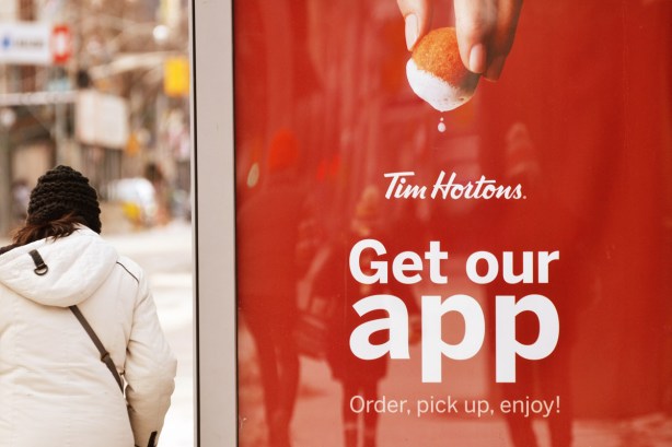 a tim horton's ad on the side of a bus shelter as a woman walks past, a timbit is being dunked into the foam of a latte 