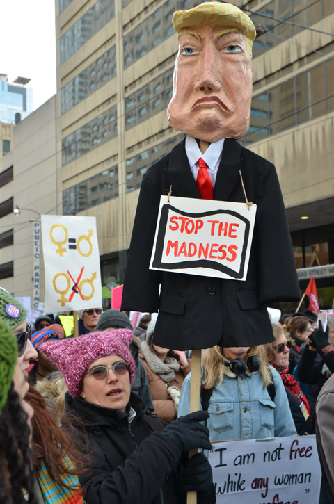 Toronto Womens march, protest parade, woman holding up an effigy of Trump with a sign that says stop the madness. Others around her walking in the protest march 