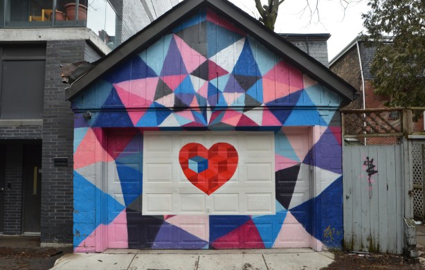 the front of a garage is covered with street art. A bright red heart is in the middle from which coloured geometric shapes eminate outwards and cover the entire front of the building. 