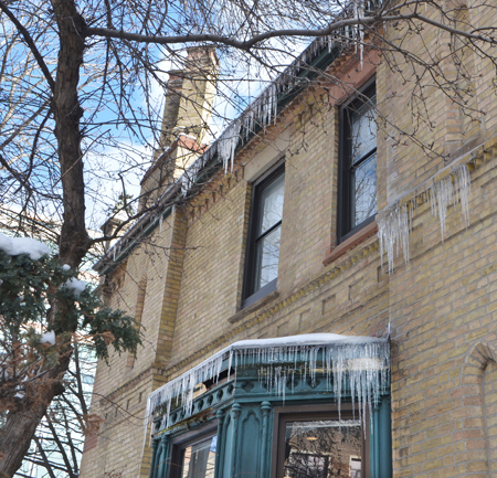 older yellowish brick building with green bay window, with icicles on the eaves of bopth roof and window 